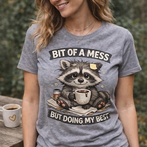 May include: Gray t-shirt featuring a cartoon raccoon holding a coffee cup, with the text "BIT OF A MESS BUT DOING MY BEST". A white coffee mug with a heart is on a wooden table.