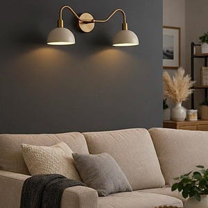May include: A wall-mounted light fixture with two cream-colored dome-shaped shades and a gold-toned frame. The fixture is mounted on a dark gray wall above a beige sofa with pillows and a wooden coffee table.
