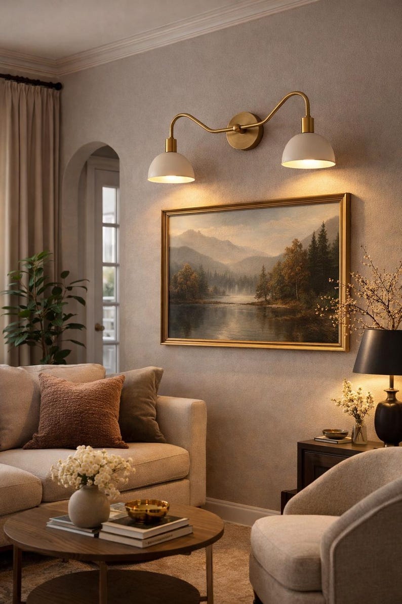 May include: A living room scene featuring a beige sofa with decorative pillows, a round wooden coffee table, and a framed landscape painting. Above the painting, a brass and white double-light fixture provides illumination. The room has a neutral color palette.