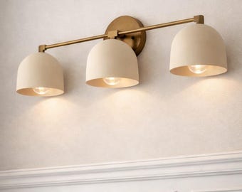 May include: A three-light vanity fixture with a brass finish and off-white dome shades. The fixture is mounted on a wall with a neutral color and white trim. Each shade houses a warm-toned light bulb.