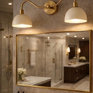 May include: A gold-toned, two-light wall sconce with off-white shades is mounted above a bathroom mirror. The mirror has a gold frame and reflects the bathroom's interior, including a marble countertop and gold fixtures.