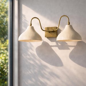 May include: A two-light wall sconce featuring cream-coloured, ribbed shades and brass-coloured curved arms. The sconce is mounted on a square brass-coloured base. The sconce is mounted on a light-coloured wall near a window.