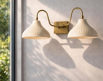 May include: A double wall sconce featuring two cream-colored, ribbed, bell-shaped shades attached to a central rectangular brass mounting plate by two curved brass arms. The fixture has a metallic finish and a classic, symmetrical design.
