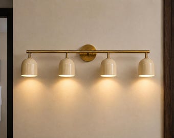 May include: A wall-mounted light fixture featuring a horizontal brass bar and a circular brass backplate. Four dome-shaped, cream-colored shades are attached to the bar, each emitting a warm, downward-facing light from the open bottom.