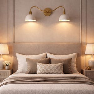 May include: A bedroom scene featuring a bed with a beige headboard, pillows, and a light brown blanket. Above the bed, a gold-toned wall-mounted light fixture with two white lampshades is installed. Two bedside lamps with white shades flank the bed.