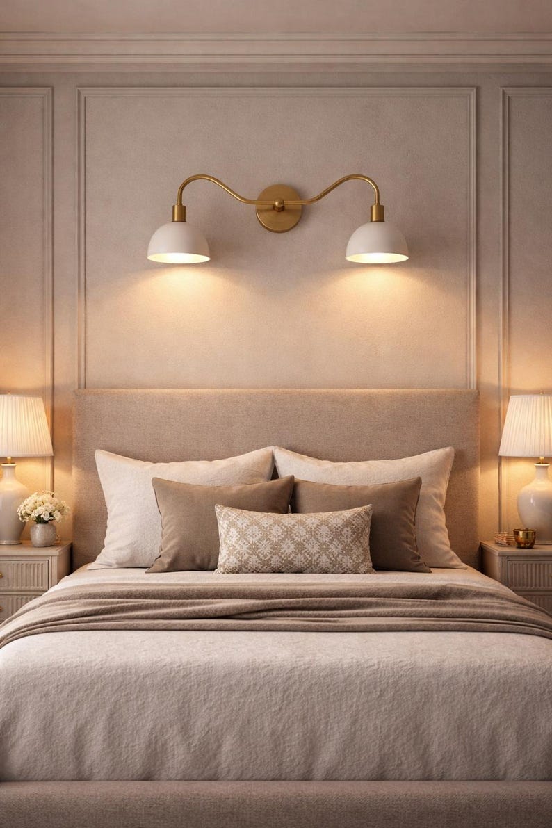May include: A bedroom scene featuring a bed with a beige headboard, pillows, and a light brown blanket. Above the bed, a gold-toned wall-mounted light fixture with two white lampshades is installed. Two bedside lamps with white shades flank the bed.