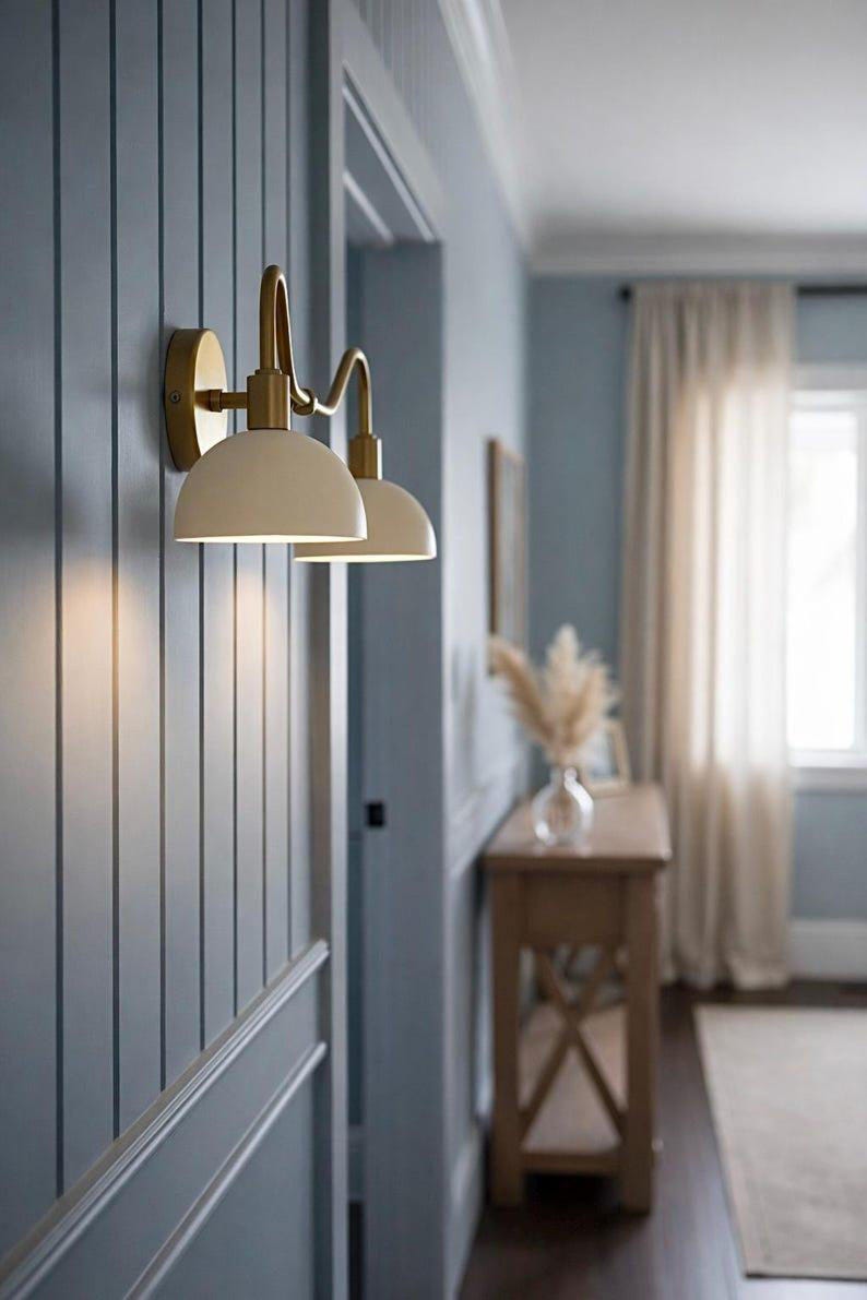 May include: Cream-colored wall sconces with a brass finish are mounted on a blue paneled wall. The sconces have a curved design and are positioned near a doorway. The room has a neutral color palette with a wooden console table.