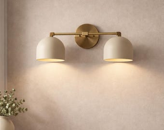 May include: A two-light vanity fixture with a brass finish and off-white dome shades. The fixture is mounted on a light-colored wall, with the lights illuminating the area below. The design is modern and minimalist.