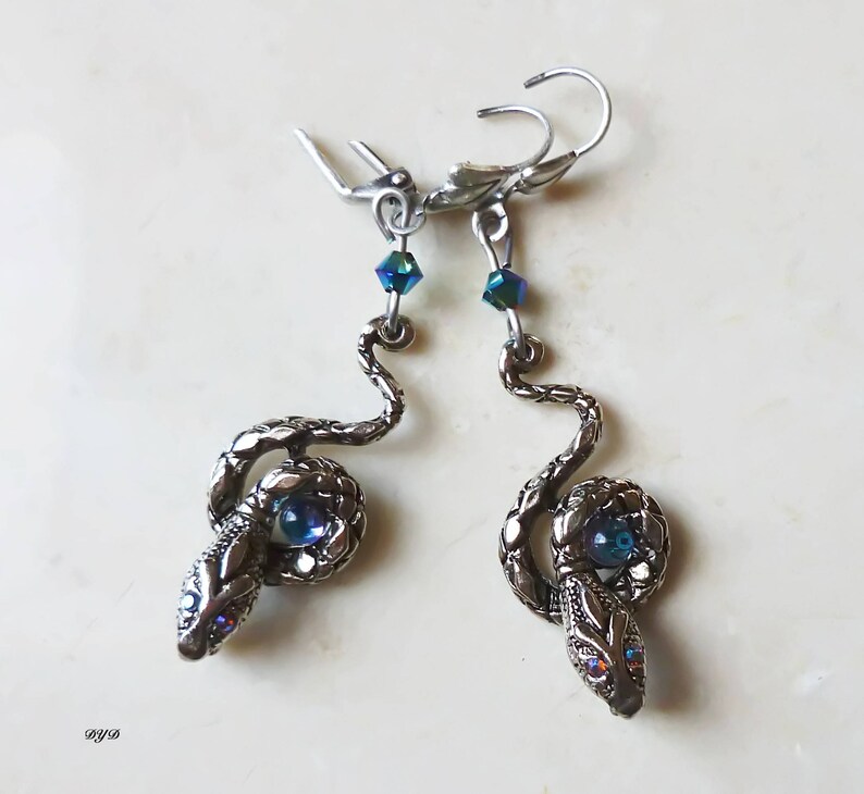 K&ouml;nnte beinhalten: Ein Paar silberne Schlangenohrringe mit blauen, irisierenden Glasaugen. Die Ohrringe h&auml;ngen und haben einen Hakenverschluss.