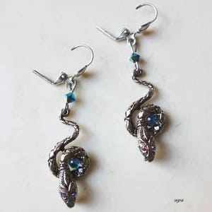 K&ouml;nnte beinhalten: Ein Paar silberne Schlangenohrringe mit blauen, schillernden Kristall-Akzenten. Die Schlangen sind zusammengerollt und haben ein blaues Kristall-Auge.