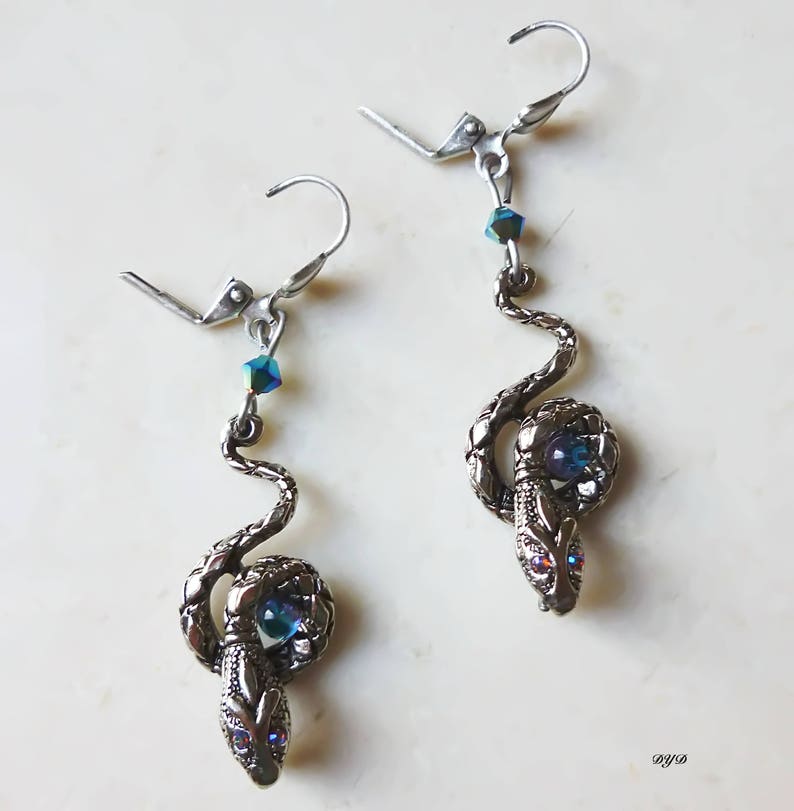 K&ouml;nnte beinhalten: Ein Paar silberne Schlangenohrringe mit blauen, schillernden Kristall-Akzenten. Die Schlangen sind zusammengerollt und haben ein blaues Kristall-Auge.