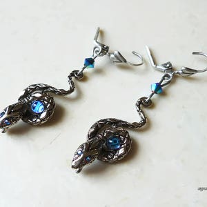 K&ouml;nnte beinhalten: Ein Paar silberne Schlangenohrringe mit blauen, schillernden Kristall-Akzenten. Die Schlangen sind gewickelt und haben ein detailliertes Design.