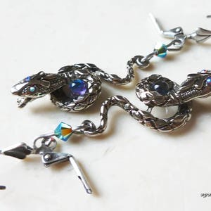 Może przedstawiać: Srebrne kolczyki w kształcie węża z niebieskimi i tęczowymi akcentami z kryształów. Węże są zwinięte i mają szczegółowe łuski. Kolczyki są w stylu wiszących z zapięciem na haczyk.