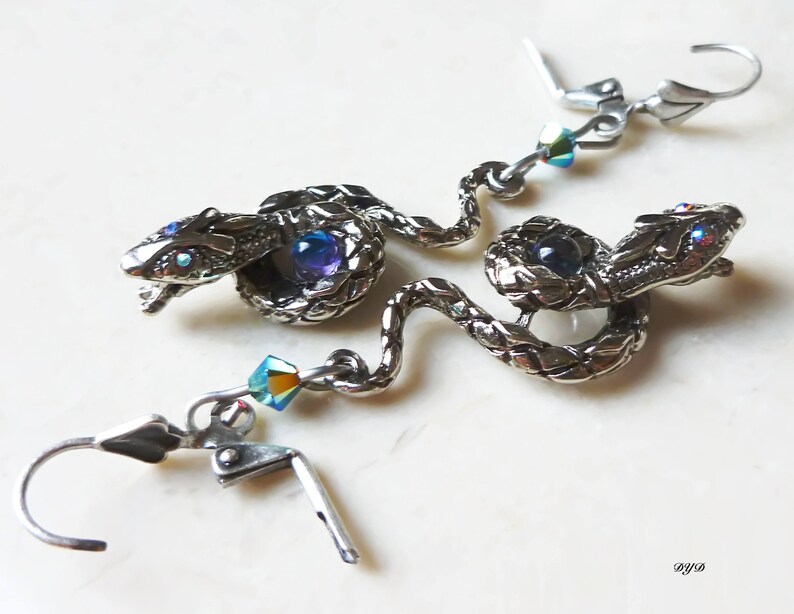 K&ouml;nnte beinhalten: Silberne Schlangenohrringe mit blauen und schillernden Kristall-Akzenten. Die Schlangen sind gewickelt und haben detaillierte Schuppen. Die Ohrringe sind im H&auml;nge-Stil mit einem Hakenverschluss.