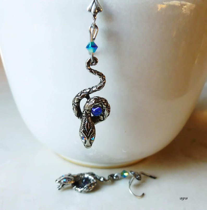 K&ouml;nnte beinhalten: Ein Paar silberne Schlangenohrringe mit blauen und schillernden Kristall-Akzenten. Die Schlange ist zusammengerollt und hat ein blaues Kristall-Auge.