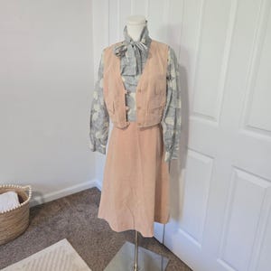 May include: A light peach-colored vest and matching skirt set paired with a long-sleeved blouse featuring a grey and white floral pattern. The blouse has a high neck with a tie detail, while the vest includes front pockets and button closures.