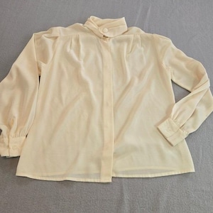 May include: A cream-colored blouse with long sleeves and a high collar. The blouse has a button-down front and buttoned cuffs. The fabric appears to be lightweight and slightly sheer, with a subtle sheen.