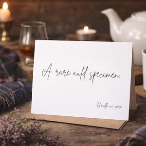 May include: A white card with the text "A rare and specimen. Handle with care" is propped up on a brown envelope. The card is surrounded by a cozy scene with a cup, teapot, candles, and a plaid blanket.
