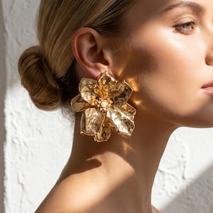 May include: A large, gold-toned metal earring shaped like a blooming flower with textured, hammered petals and a small, central white pearl accent. The earring is worn on the earlobe, showcasing a metallic finish that reflects light across its surface.