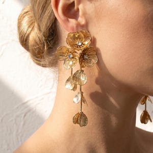 Könnte beinhalten: Ein Paar lange goldfarbene Ohrringe mit mehreren übereinanderliegenden blumenförmigen Metallblüten. Jedes Blumenelement ist durch dünne goldfarbene Ketten verbunden, und die Mitte der drei oberen Blüten ist mit einem kleinen, klaren Kristall besetzt.