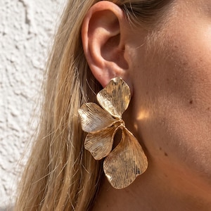 May include: A gold-toned earring featuring a cluster of three overlapping, textured petal-like shapes. The metal surface has a brushed, linear finish that catches the light, creating a layered, organic appearance that extends from the earlobe.