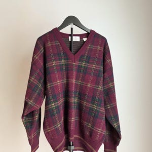 May include: A burgundy V-neck sweater with a plaid pattern in green, gold, and navy blue. The sweater is hanging on a black hanger against a white background. The sweater has long sleeves and ribbed cuffs and hem.