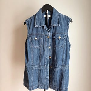 May include: A sleeveless denim vest in a medium blue wash. The vest features a collared neckline, button-down front, two chest pockets, and decorative stitching. It is hanging on a black hanger against a neutral background.
