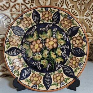 Pode incluir: Um prato decorativo de cerâmica com um design floral. O prato apresenta um centro azul escuro com flores castanhas e amarelas e folhas verdes. A borda tem um fundo creme com flores castanhas e folhas azuis. O prato é exibido em um suporte preto.