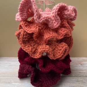 Puede incluir: Una pila de cuatro coleteros de ganchillo hechos a mano en tonos rosa, naranja y burdeos. Los coleteros están hechos con un punto texturizado, creando un efecto de volantes. Se exhiben sobre un vaso transparente.