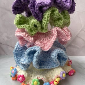 Puede incluir: Una pila de coleteros de ganchillo hechos a mano en varios colores pastel, incluyendo morado, verde, rosa y azul. El coletero inferior es de color crema y está adornado con cuentas en forma de flor. Estos accesorios para el cabello son perfectos para añadir un toque de color a cualquier peinado.