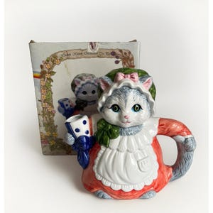 Könnte beinhalten: Eine Keramik-Teekanne in Form einer Katze, die eine grüne Haube mit rosa Schleife, eine weiße Schürze und ein rotes Kleid trägt. Die Katze hält eine blau-weiß gepunktete Tasse. Die Originalverpackung trägt den Text Kitchen Kitten Decorative Tea Pot.