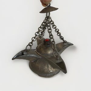 May include: A tarnished silver-toned metal oil lamp featuring three spouts and a central lid topped with a small, round red stone. The lamp is suspended by three interconnected metal chains that meet at a single top ring for hanging.