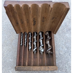 May include: A set of six metal auger drill bits stored in a wooden case with vertical slots. The wood lid features handwritten labels for sizes 1/4, 3/8, 1/2, 5/8, 3/4, and 1 inch. Each bit has a spiral flute design and a pointed tip for drilling.