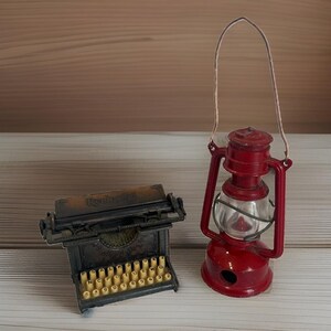 May include: A miniature dark brown typewriter with cream-colored keys and a red metal lantern with a clear glass globe and a thin wire handle. The typewriter features the word Remington embossed on the top frame above the keyboard.
