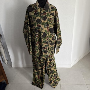 May include: A long-sleeve button-down shirt and matching trousers set featuring a multi-colored camouflage pattern in shades of dark brown, olive green, and tan. The shirt has a pointed collar and a front button closure, while the pants are loose.
