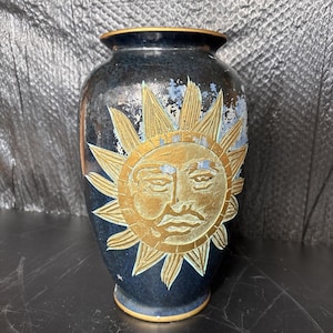 May include: A dark blue ceramic vase with a gold sun face design. The sun has a detailed face with a mustache and rays extending outward. The vase has a textured surface and a gold rim.