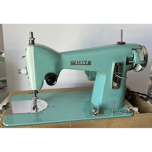 Pode incluir: Uma máquina de costura vintage verde-menta com a marca WHITE gravada em prata na lateral. A máquina possui uma placa de agulha de metal, uma alavanca de ajuste de comprimento de ponto, um volante e um suporte de carretel numa base de madeira.