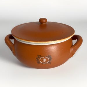 May include: A round, terracotta-colored ceramic cooking pot with two side handles and a matching lid. The pot features a white interior rim and a circular logo on the front that reads LINEA FUOCO Mamma Rd Lucca with a small floral graphic.