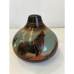 May include: A rounded ceramic vase with a narrow neck, featuring a hand-painted scene of a black wolf howling at the moon against a light blue background. The base and neck are dark brown, with orange-toned mountain silhouettes at the bottom.