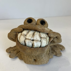 Könnte beinhalten: Eine skurrile Keramikskulptur einer Kreatur mit einem breiten, zahnigen Grinsen. Die Skulptur hat große, runde Augen und eine strukturierte, erdig-braune Oberfläche. Die Zähne sind cremefarben mit gesprenkelten Details.