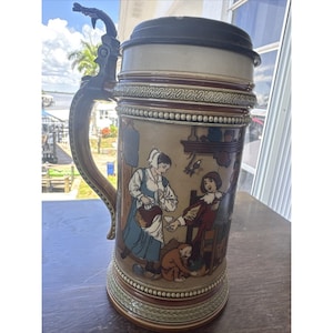May include: A tall ceramic beer stein featuring a dark brown lid and a metal handle with a dragon-shaped thumb lift. The body displays a colorful illustration of figures in historical attire within an interior setting, accented by decorative beaded bands.