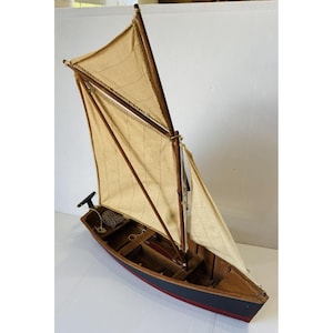 May include: A model sailboat featuring a dark blue hull with a red trim line, a wooden deck, and two beige fabric sails attached to wooden masts. The interior of the boat shows wooden benches, a small net, and a black tiller at the stern.