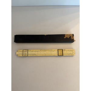 May include: A beige slide rule with two metal cursors and black printed numerical scales, accompanied by a rectangular black storage box with visible wear on the top edge. The slide rule features the text Lawrence Engineering Service.