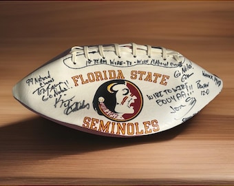 May include: A white leather football featuring the Florida State Seminoles logo in garnet and gold. The ball is covered in black ink signatures and handwritten text including '1st TEAM WIRE-TO-WIRE NATIONAL CHAMPS' and 'GO NOLES'.