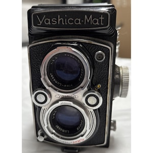 Può includere: Una fotocamera reflex a doppia lente Yashica-Mat nera. La fotocamera ha due obiettivi, un obiettivo superiore con la scritta "View-Lumaxar" e un obiettivo inferiore con la scritta "Lumaxar". C'è anche la scritta "Yashica-Mat" in alto.