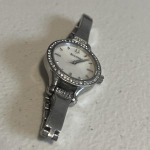 Puede incluir: Un reloj de pulsera Bulova plateado con esfera blanca y bisel con pequeñas piedras transparentes. El reloj tiene una correa plateada y un diseño clásico y elegante. La esfera muestra el logotipo de Bulova.