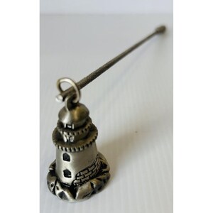 May include: A silver-toned metal lighthouse charm attached to a long, thin, textured metal rod. The lighthouse features a domed top, a balcony, and a base with a brick-patterned section. The entire object is crafted from a metallic material.