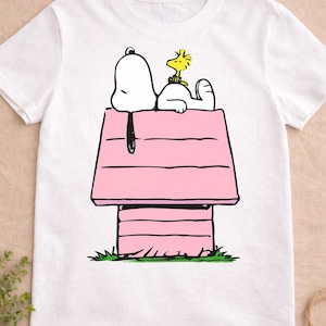 May include: White t-shirt featuring a cartoon illustration of Snoopy and Woodstock. Snoopy is lying on top of his pink doghouse, with Woodstock resting on his back. The design is set against a white background.