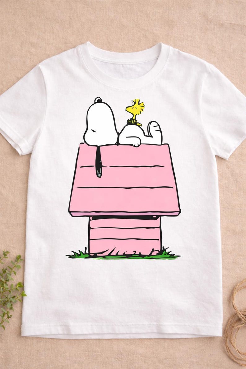 May include: White t-shirt featuring a cartoon illustration of Snoopy and Woodstock. Snoopy is lying on top of his pink doghouse, with Woodstock resting on his back. The design is set against a white background.