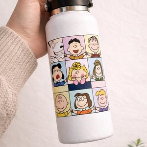 May include: White insulated water bottle featuring a colorful cartoon panel design with various characters. The bottle has a black handle and lid. A fun and functional item for carrying beverages.